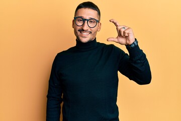 Handsome man with tattoos wearing turtleneck sweater and glasses smiling and confident gesturing with hand doing small size sign with fingers looking and the camera. measure concept.