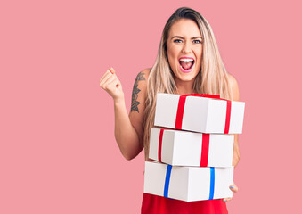 Young beautiful blonde woman holding birthday gifts screaming proud, celebrating victory and success very excited with raised arms