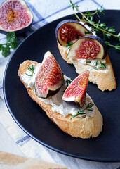 Two sandwiches with goat cheese, fig and thyme on fresh crispy bread served on a black plate on checked white and blue tablecloth