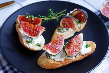 Three sandwiches with goat cheese, fig and thyme on fresh crispy bread served on a black plate on light grey background, with checked white and blue tablecloth