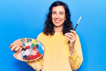 Fototapeta premium Young beautiful hispanic woman holding paintbrush and palette looking positive and happy standing and smiling with a confident smile showing teeth