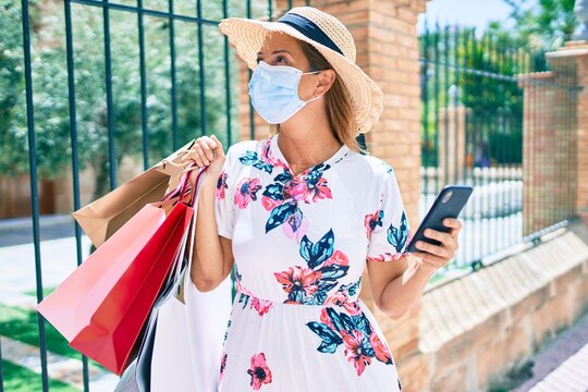 Middle age woman wearing medical mask holding shopping bags and smartphone at the city