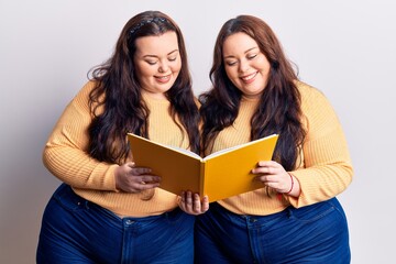 Young plus size twins holding book looking positive and happy standing and smiling with a confident smile showing teeth