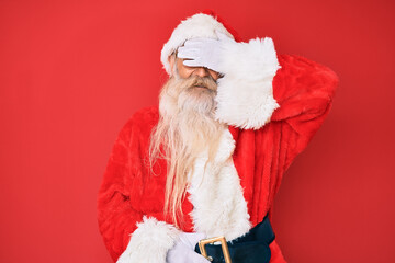 Old senior man with grey hair and long beard wearing traditional santa claus costume smiling and laughing with hand on face covering eyes for surprise. blind concept.