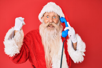 Old senior man wearing santa claus costume using vintage telephone screaming proud, celebrating victory and success very excited with raised arms