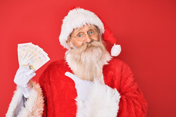 Old senior man with grey hair and long beard wearing santa claus costume holding pounds smiling happy pointing with hand and finger