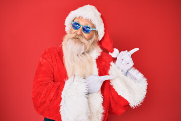 Old senior man wearing santa claus costume and sunglasses smiling and looking at the camera pointing with two hands and fingers to the side.