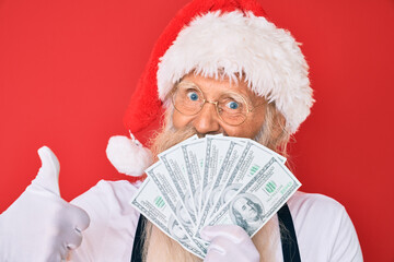 Old senior man with grey hair and long beard wearing santa claus costume holding dollars smiling happy and positive, thumb up doing excellent and approval sign