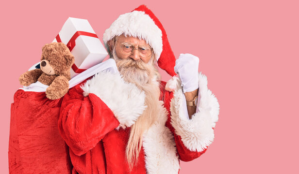 Old senior man with grey hair and long beard wearing santa claus costume holding bag with presents annoyed and frustrated shouting with anger, yelling crazy with anger and hand raised