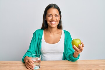 Beautiful hispanic woman drinking glass of water eating heatlhy apple looking positive and happy standing and smiling with a confident smile showing teeth