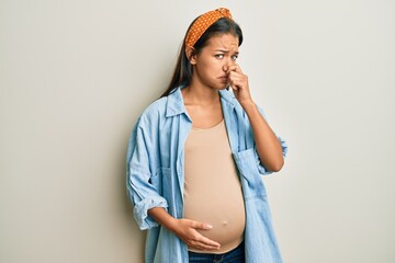 Beautiful hispanic woman expecting a baby, touching pregnant belly smelling something stinky and disgusting, intolerable smell, holding breath with fingers on nose. bad smell