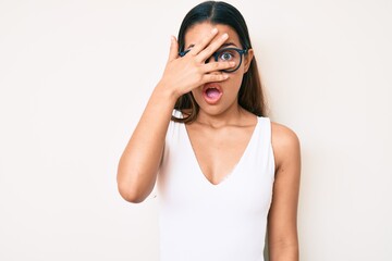 Young beautiful latin girl wearing casual clothes and glasses peeking in shock covering face and...