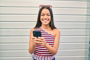 Young latin girl smiling happy using smartphone at the city.