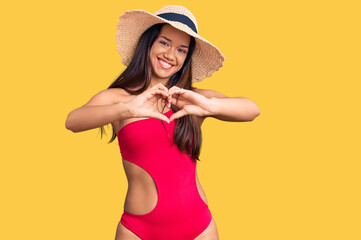 Young beautiful latin girl wearing swimwear and summer hat smiling in love doing heart symbol shape with hands. romantic concept.