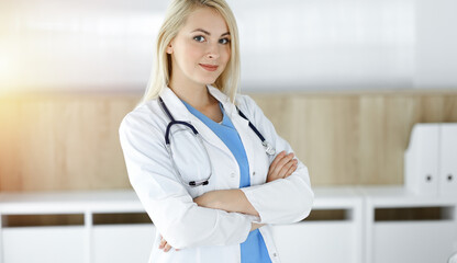 Woman-doctor at work while standing straight in sunny clinic. Blonde cheerful physician ready to help patients