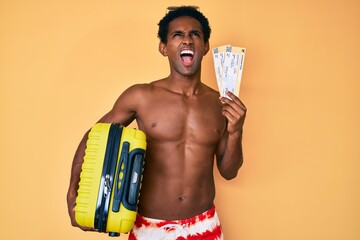 Fototapeta premium African handsome man going on summer trip holding boarding pass angry and mad screaming frustrated and furious, shouting with anger looking up.
