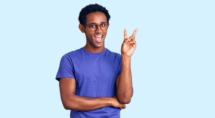 Naklejka premium African handsome man wearing casual clothes and glasses smiling with happy face winking at the camera doing victory sign. number two.