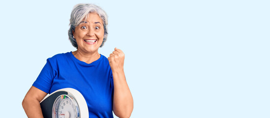 Senior woman with gray hair holding weight machine to balance weight loss screaming proud,...