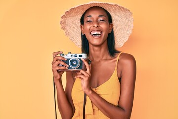 Young african american woman wearing summer hat holding vintage camera smiling and laughing hard out loud because funny crazy joke.