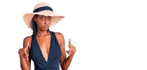 Young african american woman wearing swimsuit and summer hat pointing down looking sad and upset, indicating direction with fingers, unhappy and depressed.