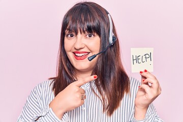 Young plus size woman wearing call center agent headset holding help reminder smiling happy...