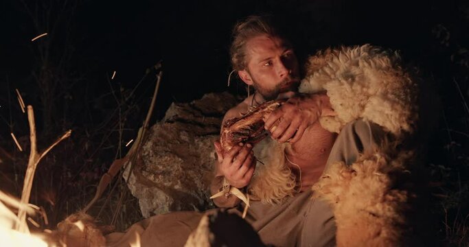 Primitive hunter gather wearing sheepskin squatting eating meat by the light of the flames of a fire at night tearing the meat from the bone with his teeth - wild caveman