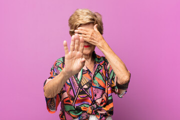 old pretty woman covering face with hand and putting other hand up front to stop camera, refusing...