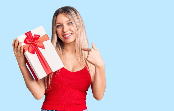 Young beautiful blonde woman holding gift pointing finger to one self smiling happy and proud