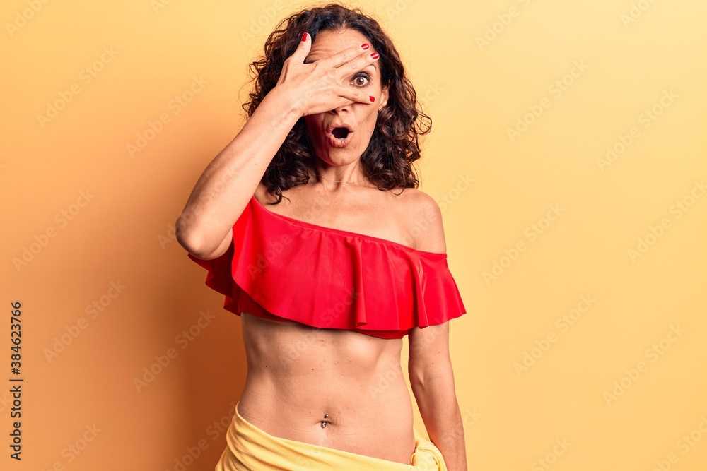 Wall mural middle age beautiful woman wearing bikini peeking in shock covering face and eyes with hand, looking