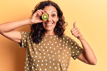Middle age beautiful woman holding kiwi over eye smiling happy and positive, thumb up doing excellent and approval sign