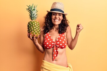 Middle age beautiful woman wearing bikini and summer hat holding pineappel screaming proud, celebrating victory and success very excited with raised arm