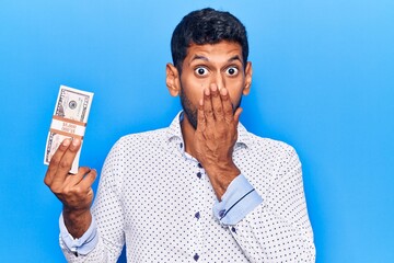 Young latin man holding dollars covering mouth with hand, shocked and afraid for mistake. surprised expression