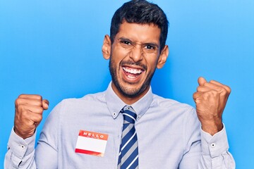 Young latin man wearing sticker with hello my name is message screaming proud, celebrating victory and success very excited with raised arms