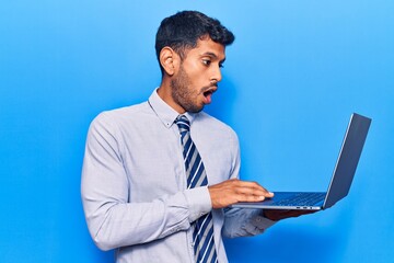 Young latin man holding laptop scared and amazed with open mouth for surprise, disbelief face
