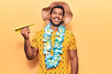 Young latin man wearing summer hat and hawaiian lei holding paper airplane looking positive and...