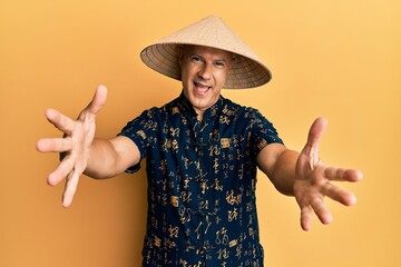 Middle age bald man wearing traditional asian straw hat looking at the camera smiling with open arms for hug. cheerful expression embracing happiness.
