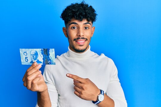 Young arab handsome man holding 5 canadian dollars banknote smiling happy pointing with hand and finger