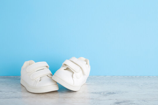 New White Sport Shoes For Little Kid On Wooden Floor At Light Blue Wall Background. Pastel Color. Baby Footwear. Empty Place For Text. Front View. Closeup.