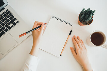 Blank paper note in female hand for any idea on desk work. Hand writting with pens to blank notebook. Planning notes writing concepts. Keyboard, coffee, cactus, blank notebook and laptop concept. 