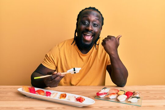 Handsome young black man eating sushi sitting on the table pointing thumb up to the side smiling happy with open mouth