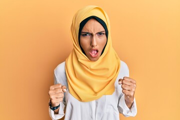 Young brunette arab woman wearing traditional islamic hijab scarf angry and mad raising fists...