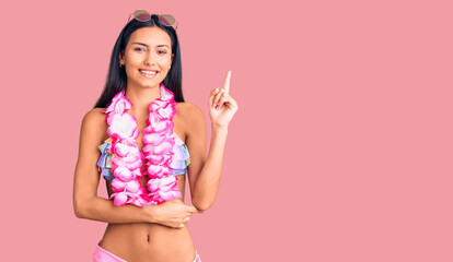 Young beautiful latin girl wearing bikini and hawaiian lei smiling happy pointing with hand and...