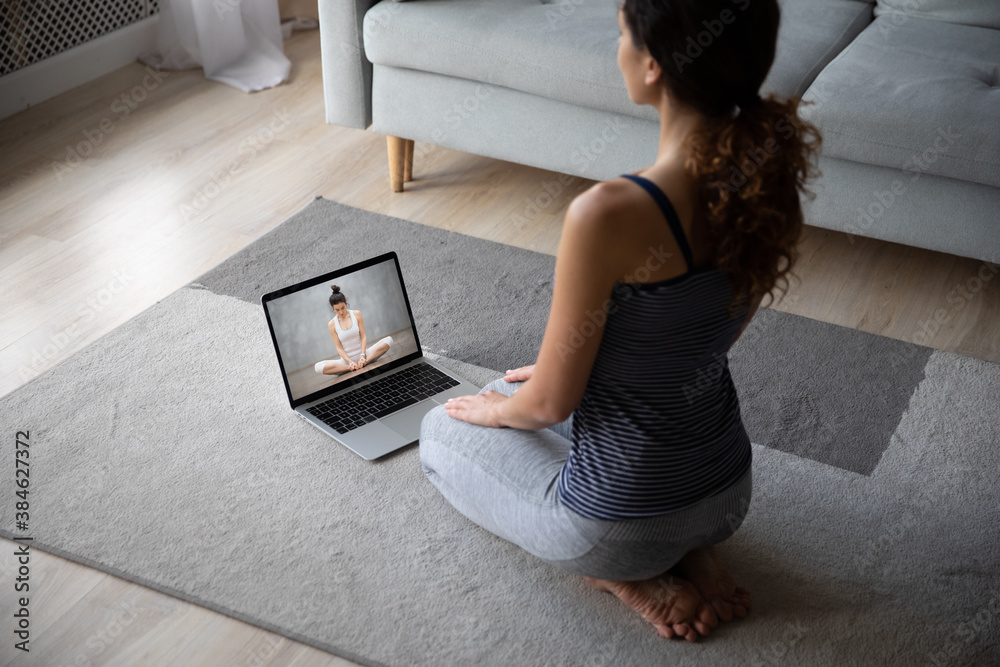 Wall mural young female sit on floor at home practice yoga mediate watching video on computer. woman do stretch