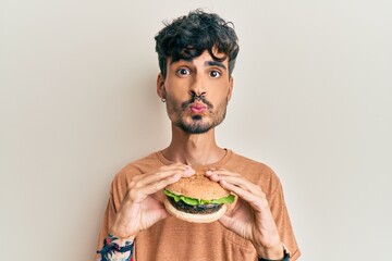 Young hispanic man eating a tasty classic burger looking at the camera blowing a kiss being lovely and sexy. love expression.