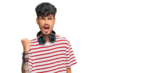 Young hispanic man listening to music using headphones angry and mad raising fist frustrated and furious while shouting with anger. rage and aggressive concept.