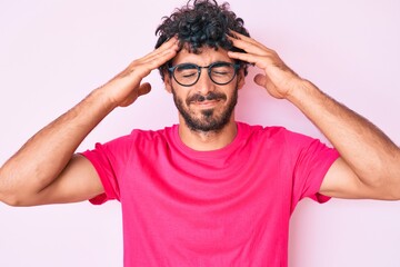 Handsome young man with curly hair and bear wearing casual clothes and glasses suffering from headache desperate and stressed because pain and migraine. hands on head.