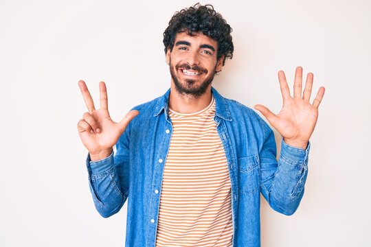 Handsome young man with curly hair and bear wearing casual denim jacket showing and pointing up with fingers number eight while smiling confident and happy.