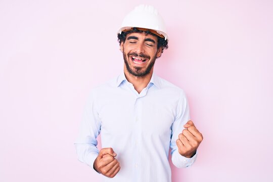 Handsome young man with curly hair and bear wearing architect hardhat celebrating surprised and amazed for success with arms raised and eyes closed