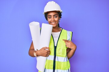 Young african american woman with braids wearing safety helmet holding blueprints smiling happy pointing with hand and finger