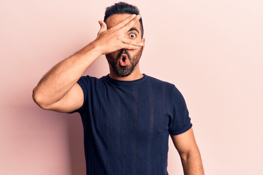 Young hispanic man wearing casual clothes peeking in shock covering face and eyes with hand, looking through fingers afraid